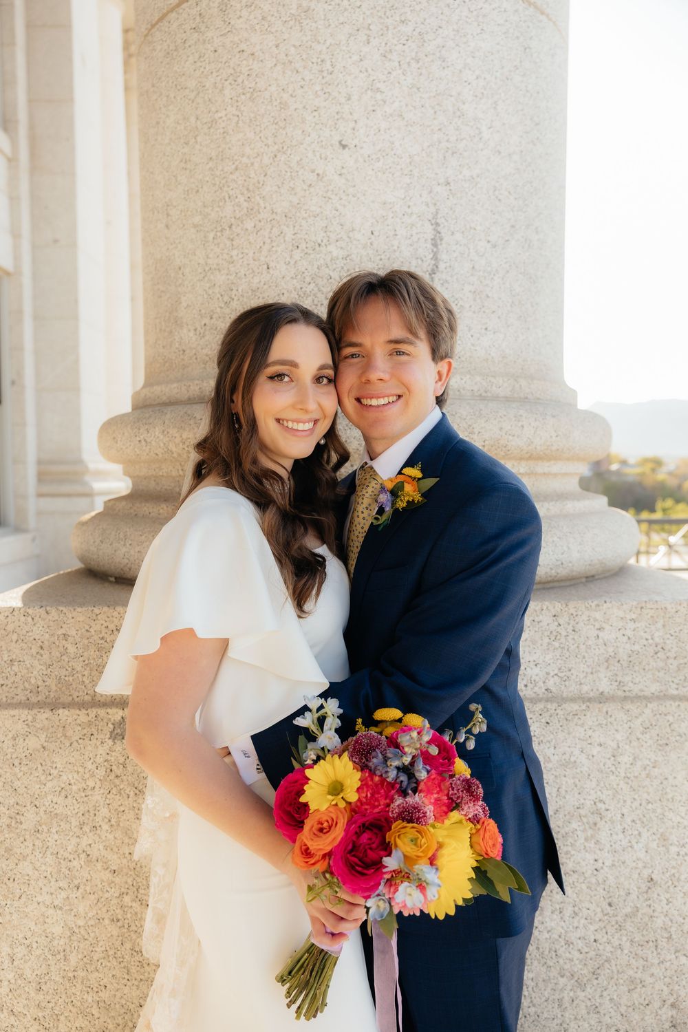 Smiling bride and groom pose by stone column, bride holding vibrant colorful bouquet of roses and gerbera daisies.