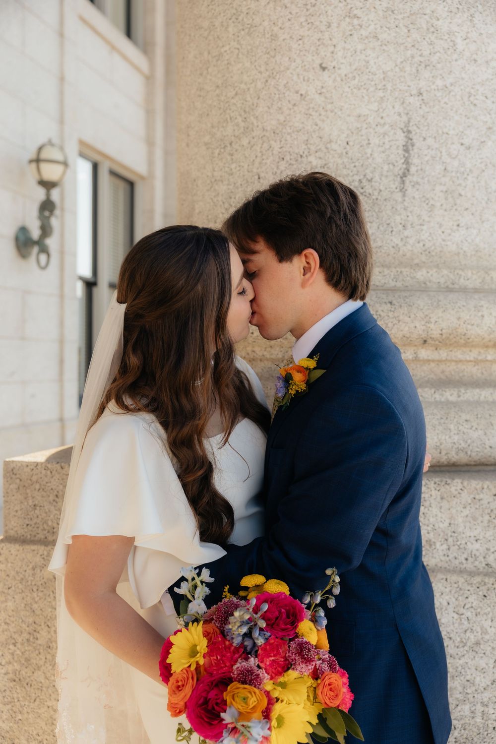 Newlyweds share a kiss outdoors, bride holding vibrant colorful bouquet in white flutter-sleeve dress with veil.