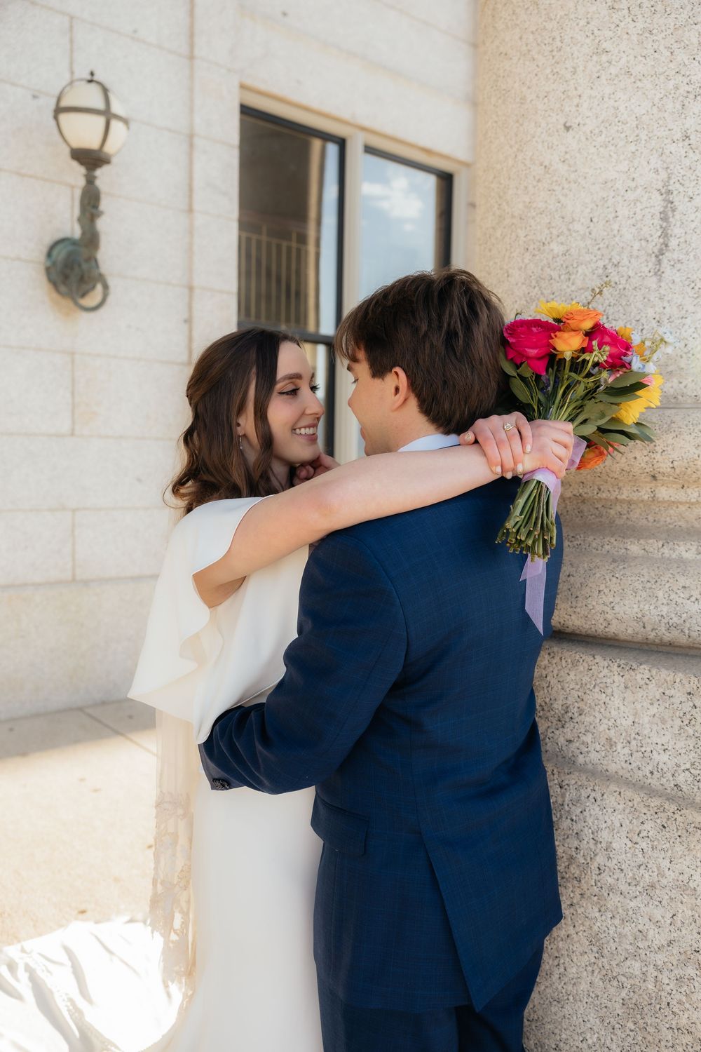 Bride and groom embrace near stone columns, she smiles holding a vibrant bouquet of red, orange, and yellow flowers.