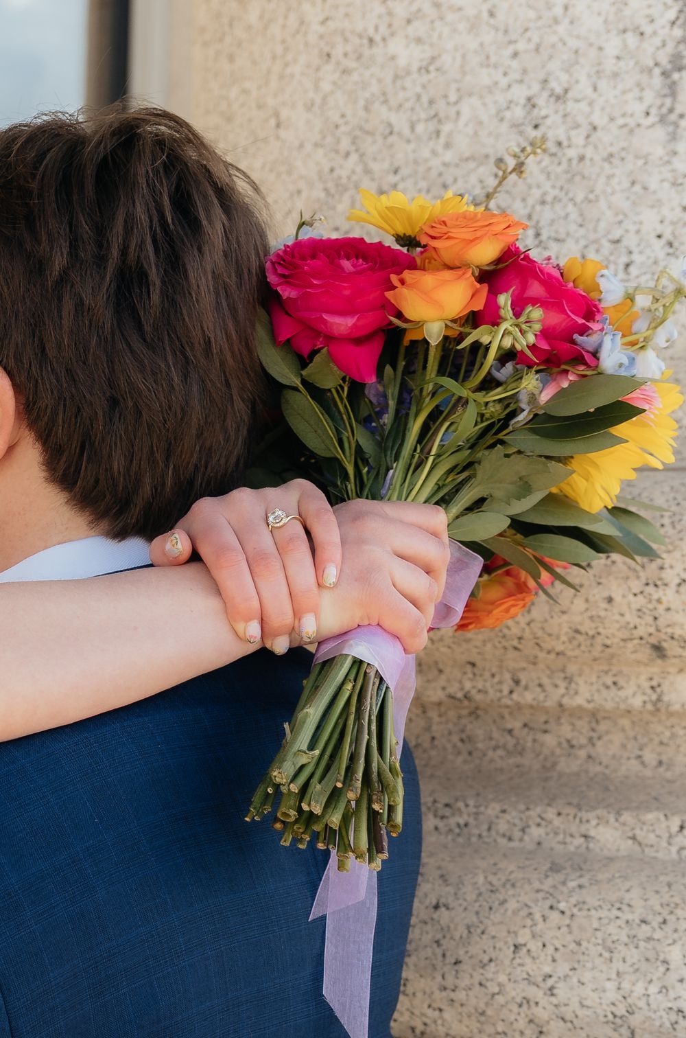 Bride holding colorful bouquet of pink, orange, and yellow flowers while embracing groom in navy suit.
