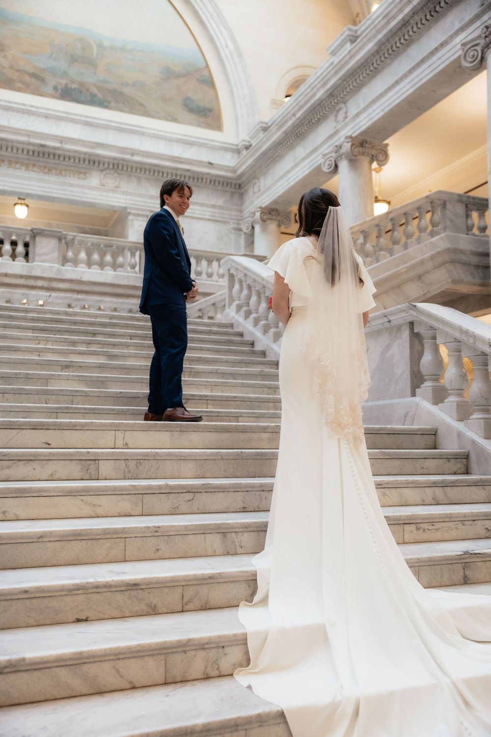 Bride in white gown with veil walks up marble staircase toward groom in navy suit at elegant wedding venue.