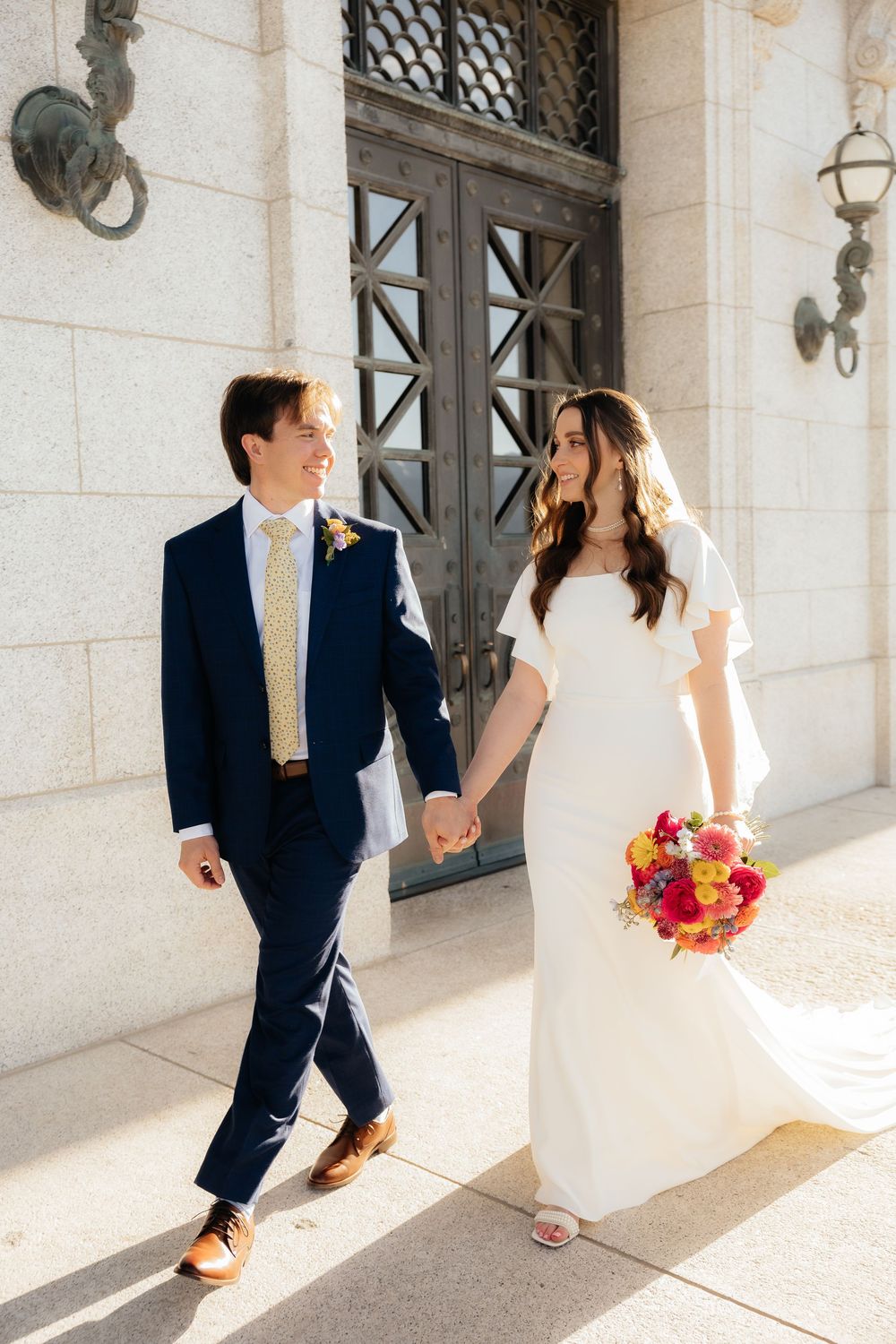 Bride and groom walk hand in hand outside a grand building, she holds a vibrant bouquet as he wears a navy suit.