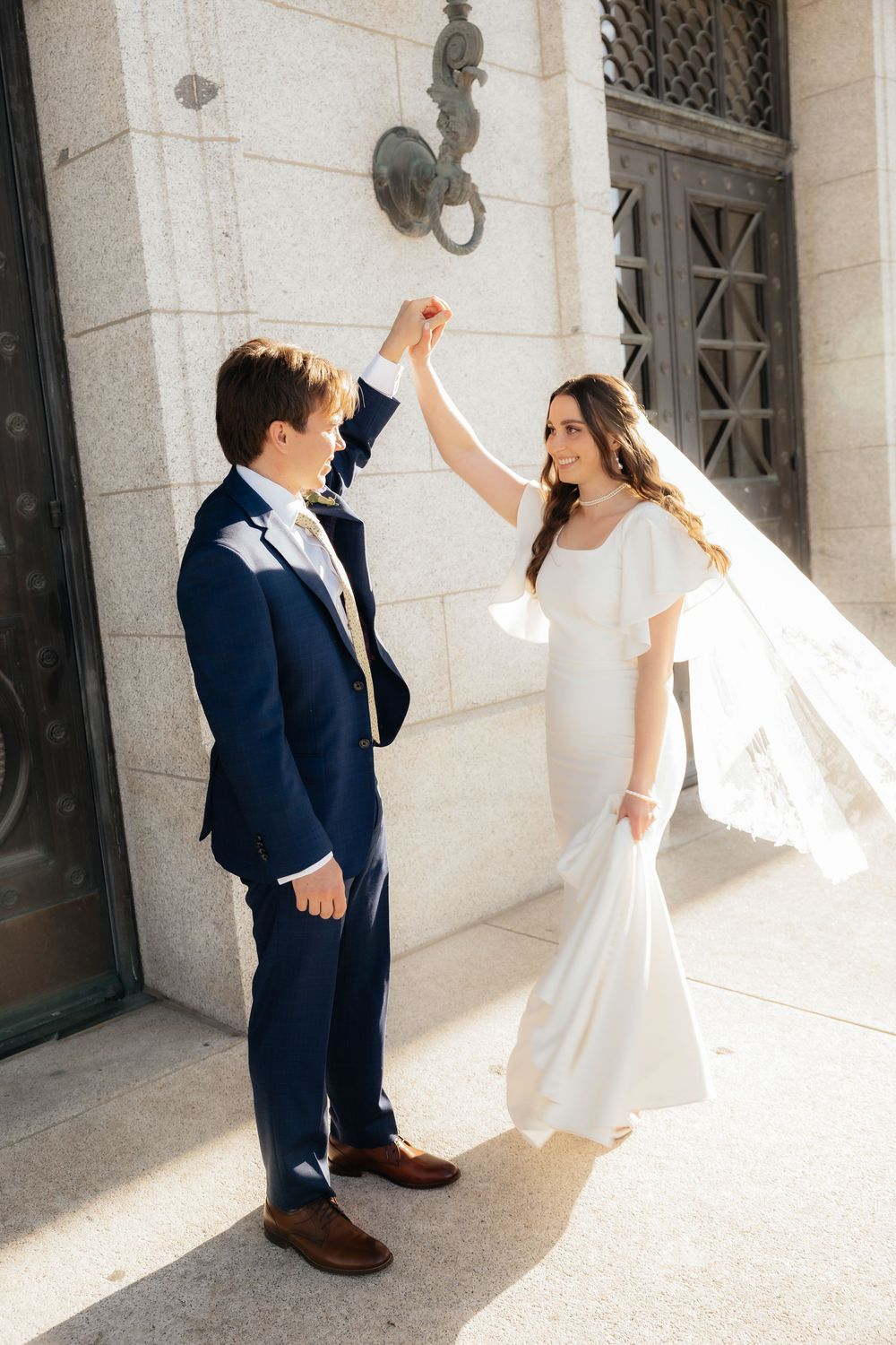 Bride and groom dancing outside a grand building, bride in white mermaid gown with veil, groom in navy suit.