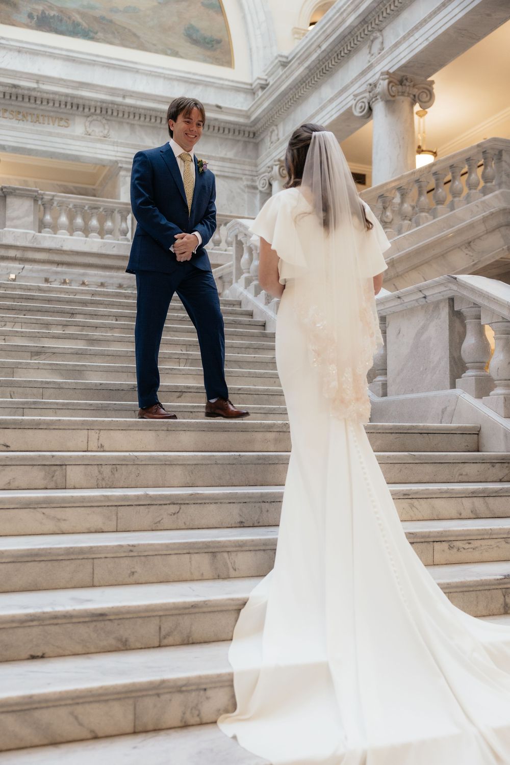 Bride and groom share a romantic moment on marble staircase, bride in elegant white gown with veil, groom in navy suit.