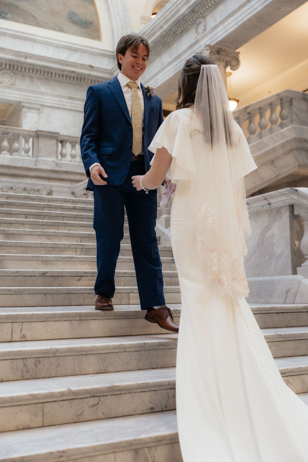 Bride and groom holding hands on marble staircase inside elegant wedding venue.