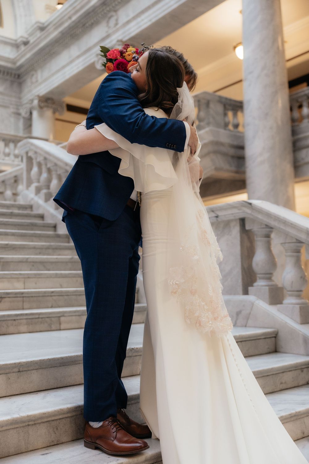 Bride and groom share a romantic embrace on marble stairs, bride in elegant white gown with floral hair piece.