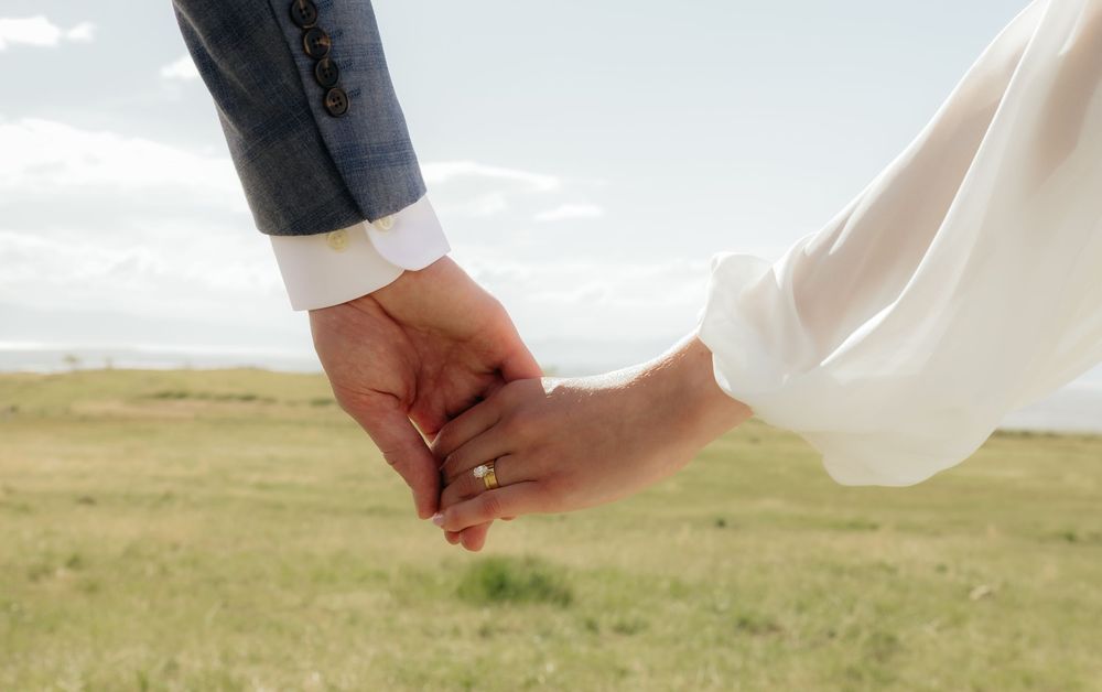 Close-up of newlyweds holding hands in a green field, bride wearing a gold wedding ring and white lace sleeve.