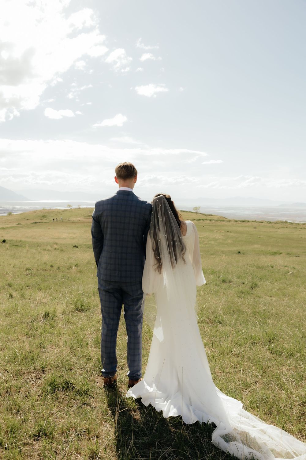 Bride and groom standing back-to-back gazing at a scenic landscape, brides long veil flowing in the breeze.