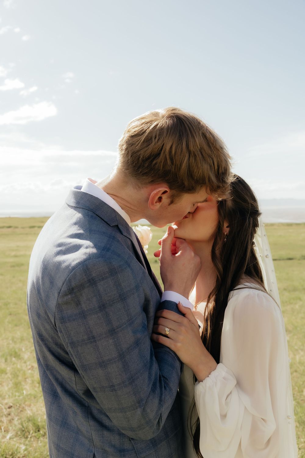 Bride and groom share a tender kiss outdoors in sunlit green field on their wedding day.
