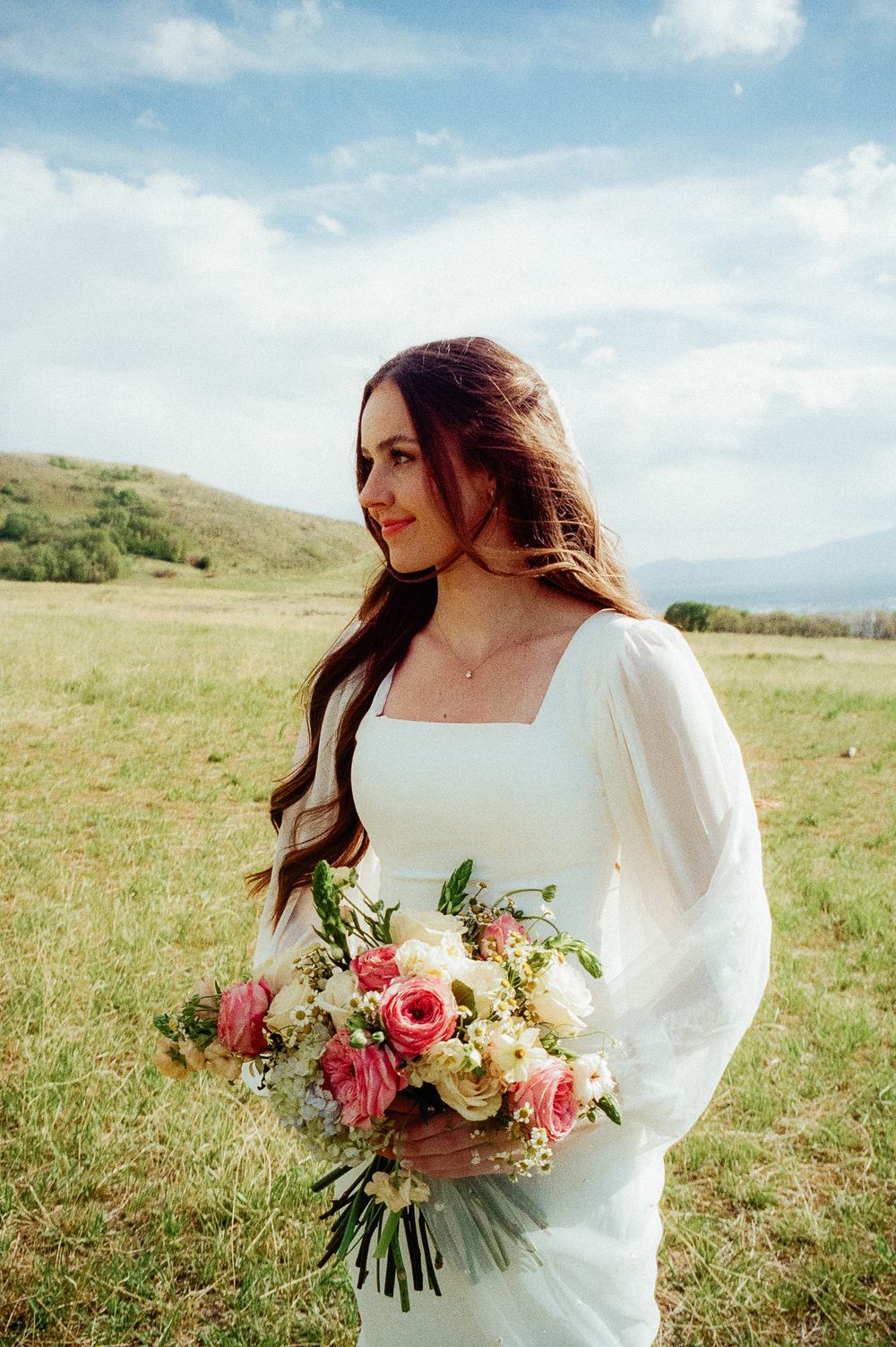 Bride in white long-sleeve gown holds pink and white bouquet in a green meadow.