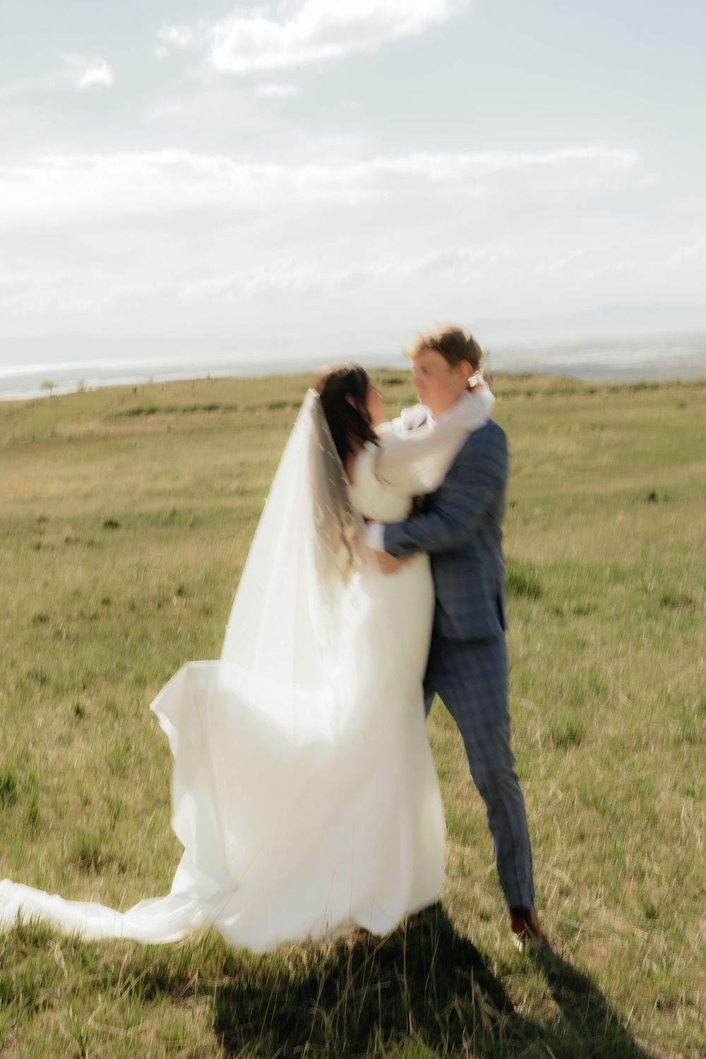 Bride and groom embrace romantically in sunlit green meadow, wedding veil billowing softly in breeze.