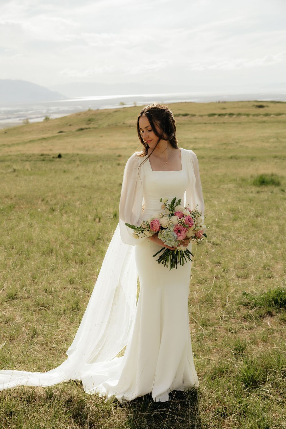 Elegant bride in a white long-sleeve gown with train holds a pink and white bouquet while standing in a scenic field.