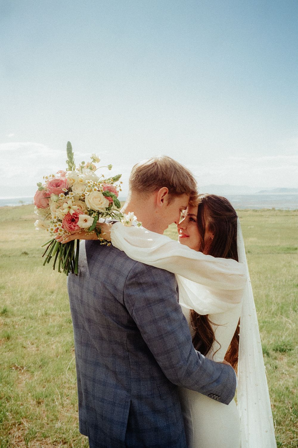Newlyweds sharing an intimate moment, groom in plaid suit embracing bride wrapped in veil holding a pink and white bouquet.