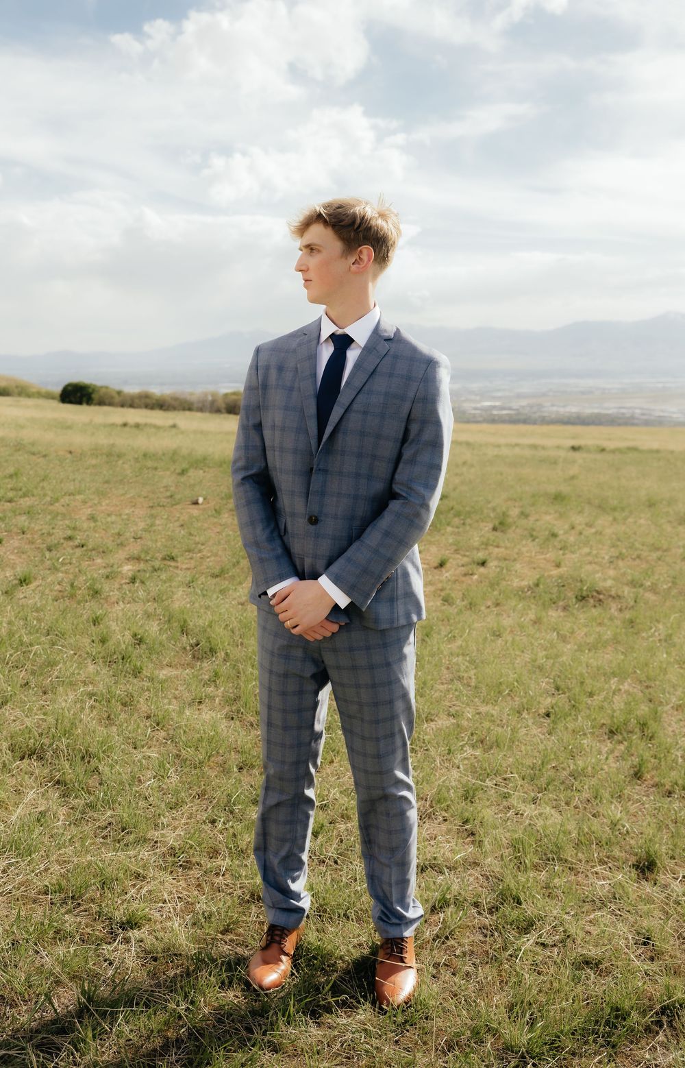 Young man in gray plaid suit with navy tie stands in open field with mountain landscape backdrop.