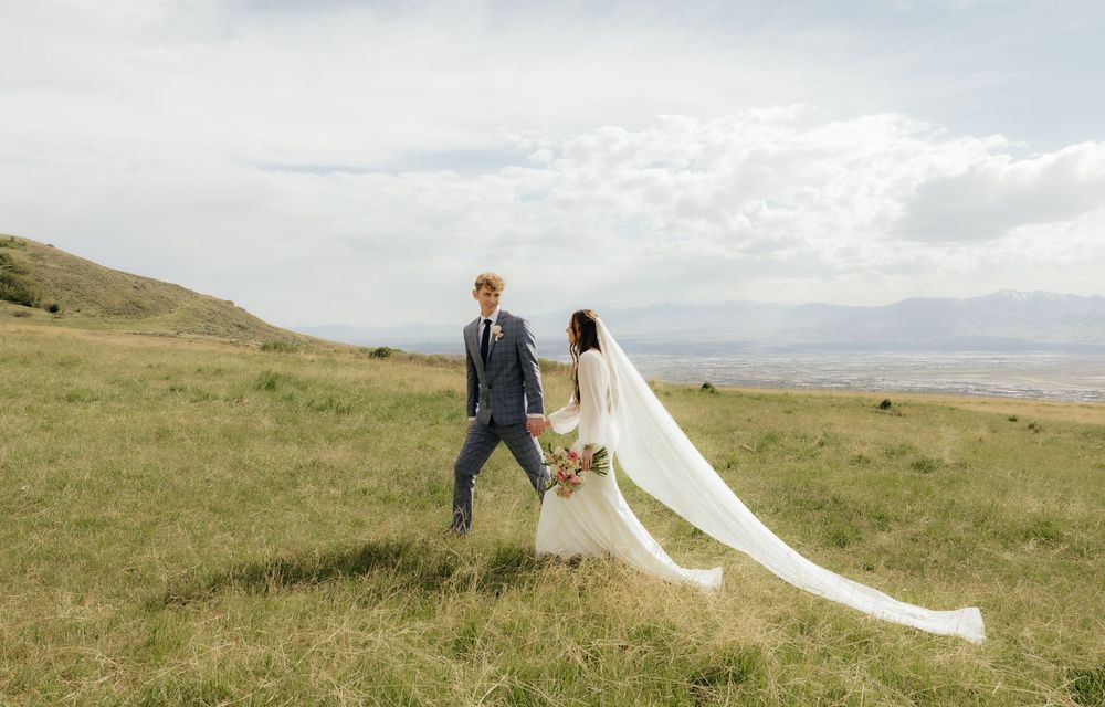 Bride and groom walk hand in hand through lush green field, long wedding veil trailing behind.