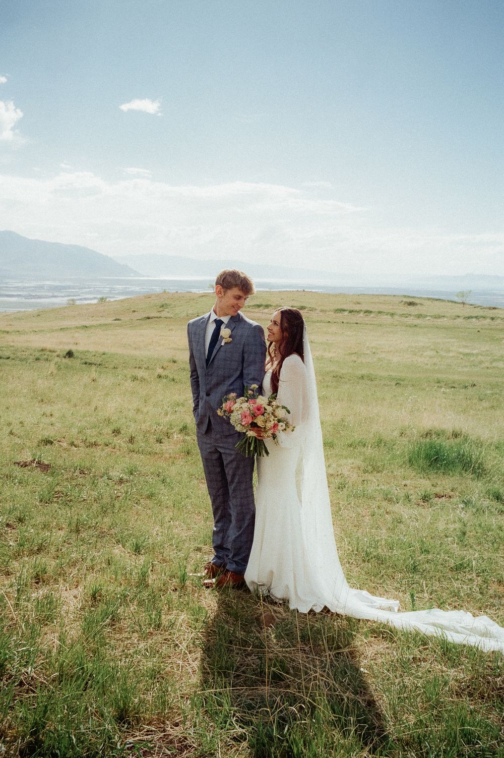 Bride and groom share intimate glance in expansive green field with mountain and lake views behind them.