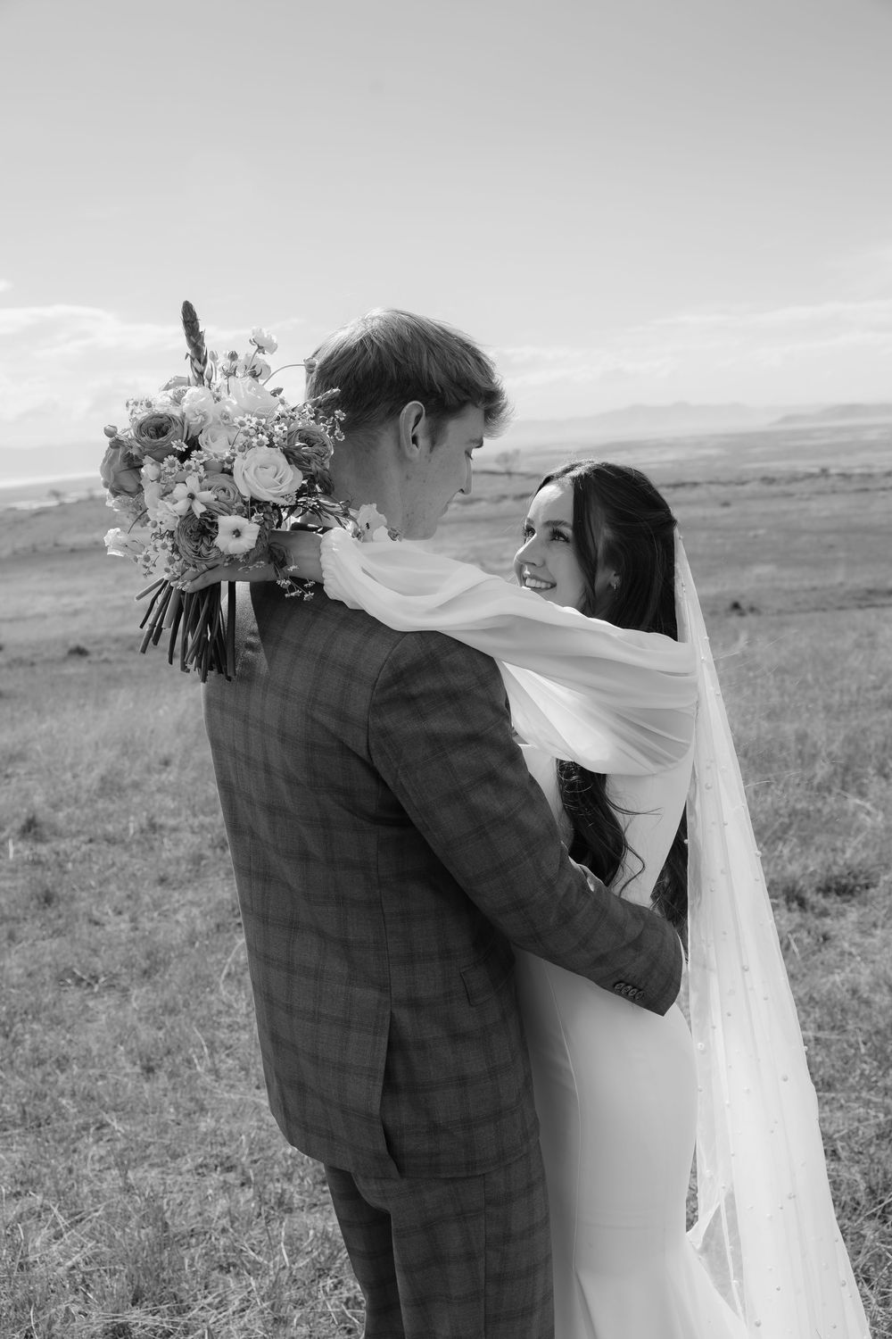 Black and white photo of bride and groom embracing in a field, bride smiling over grooms shoulder holding bouquet.