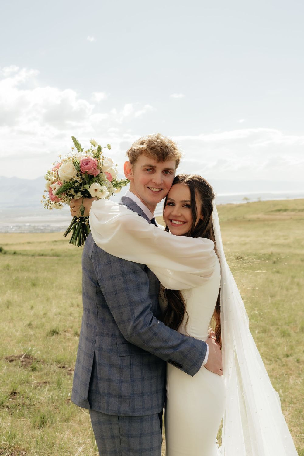Bride and groom embracing in a green field, she holds pink bouquet, both smiling, mountains in background.
