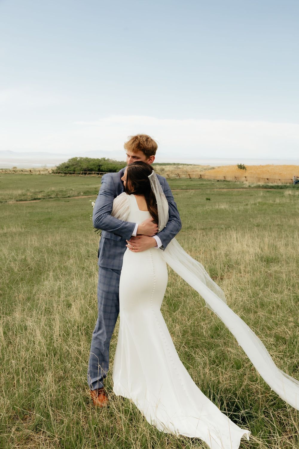 Bride and groom embracing in a green field, bride in white gown with flowing veil, groom in blue plaid suit.