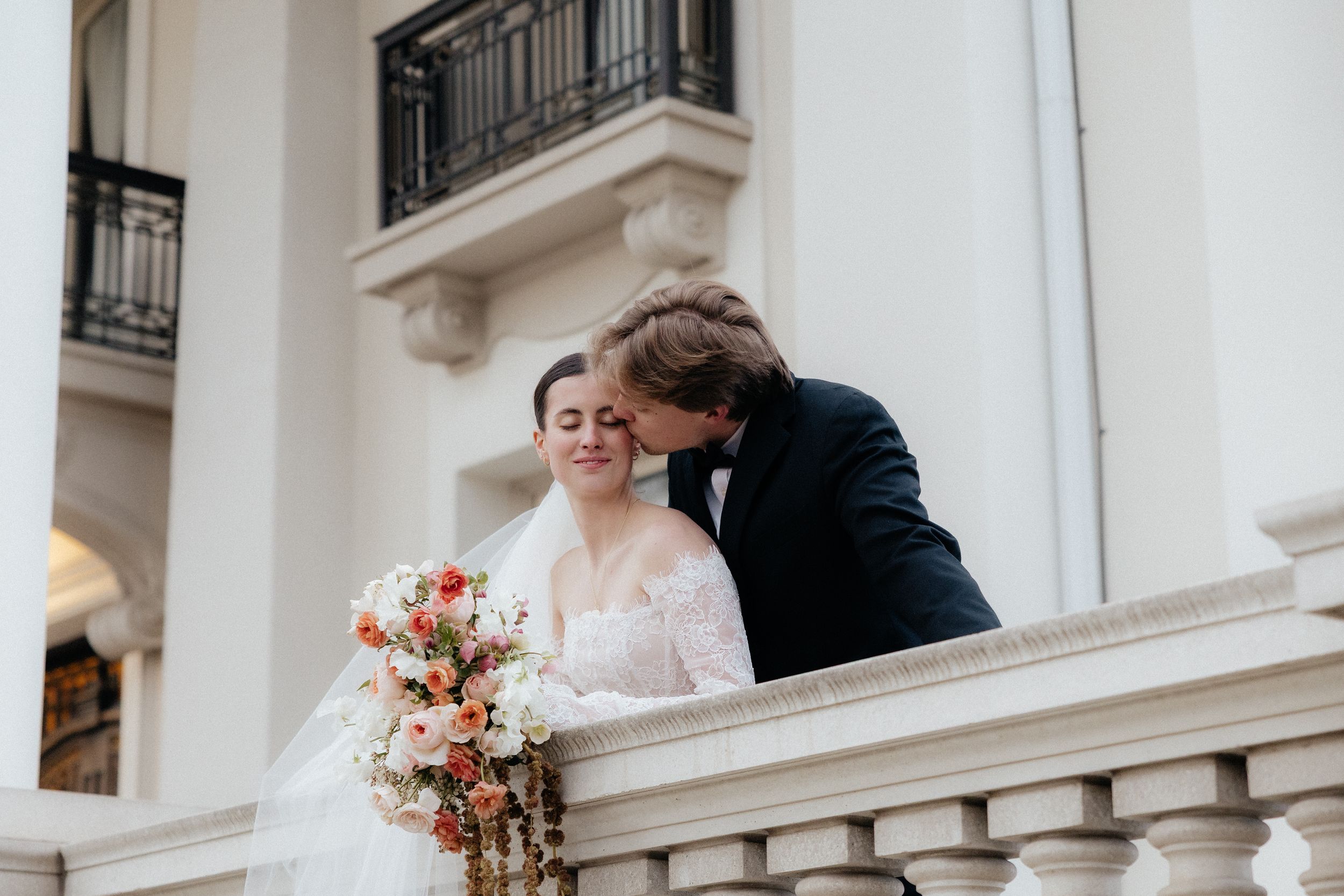 Groom kissing bride on balcony, bride holding colorful bouquet in elegant white wedding attire.