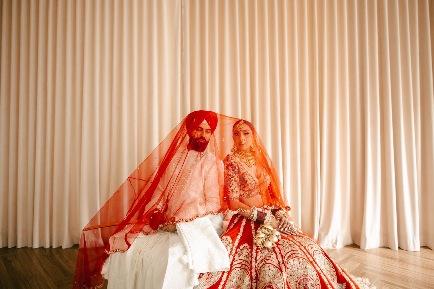 Sikh bride and groom in traditional red and orange wedding attire seated together against a cream curtain backdrop.