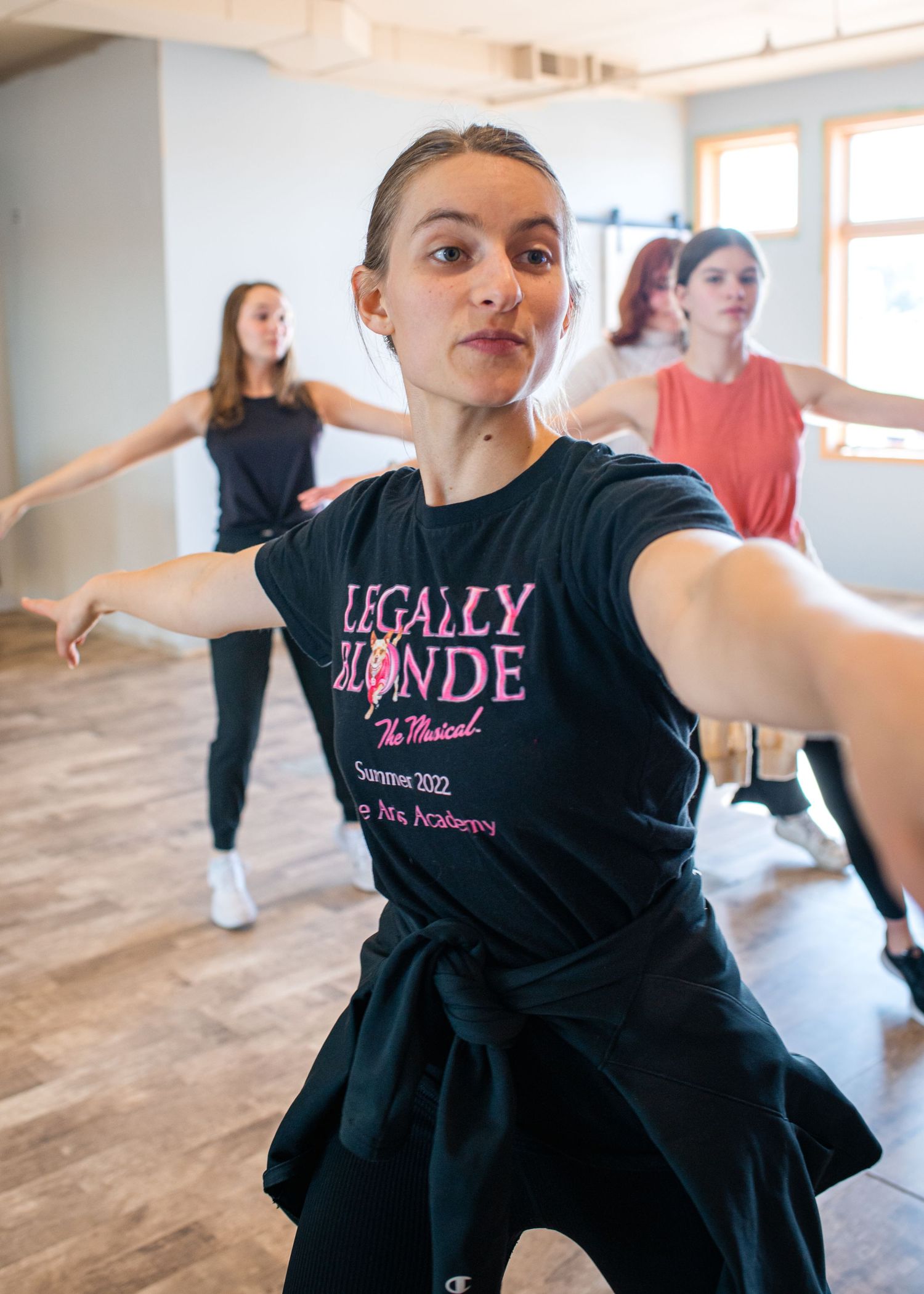 Young dancer in Legally Blonde the Musical t-shirt rehearsing choreography with other dancers in a bright studio.