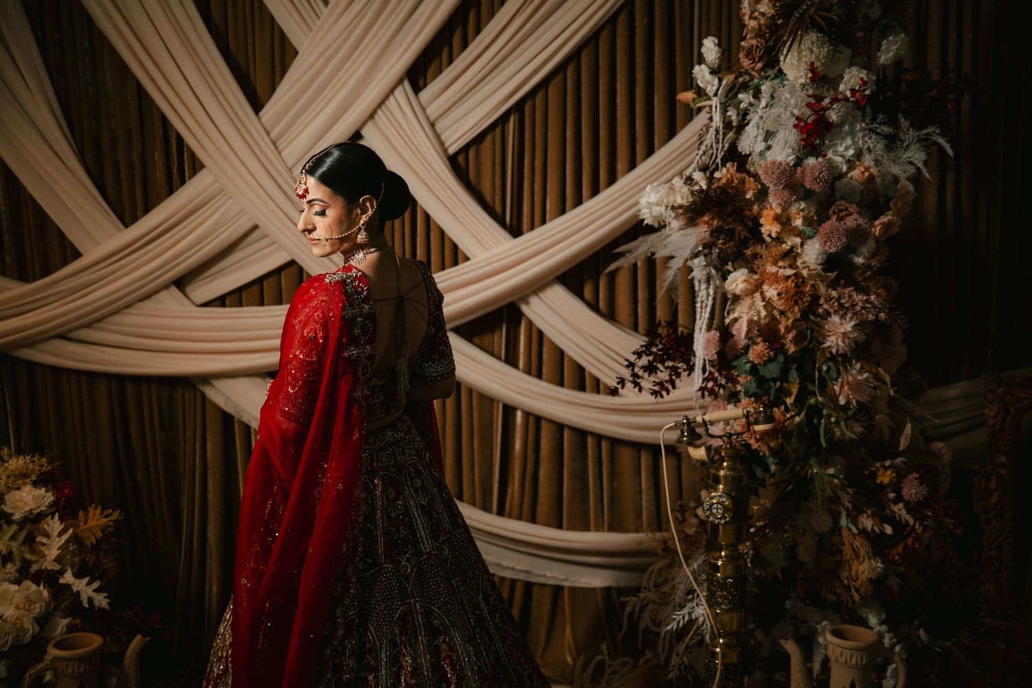 Bride in red embroidered lehenga poses before draped fabric backdrop with floral arrangement.