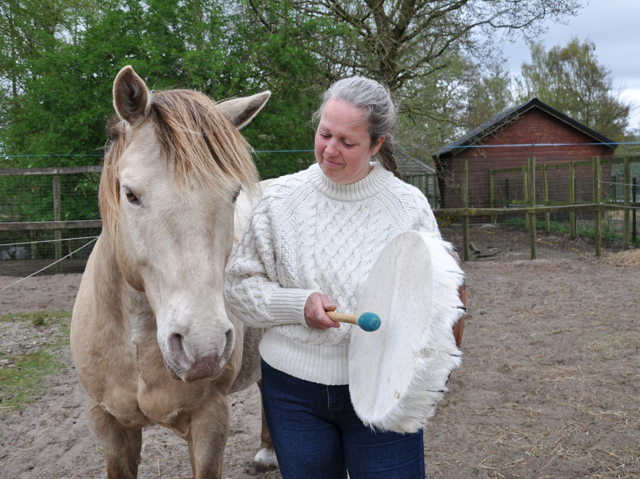 Woman in white sweater holds a large feather fan beside a palomino horse in a paddock.