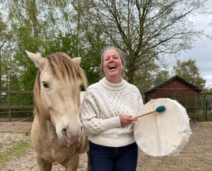 Laughing woman in white sweater holds a frame drum beside a palomino horse outdoors.