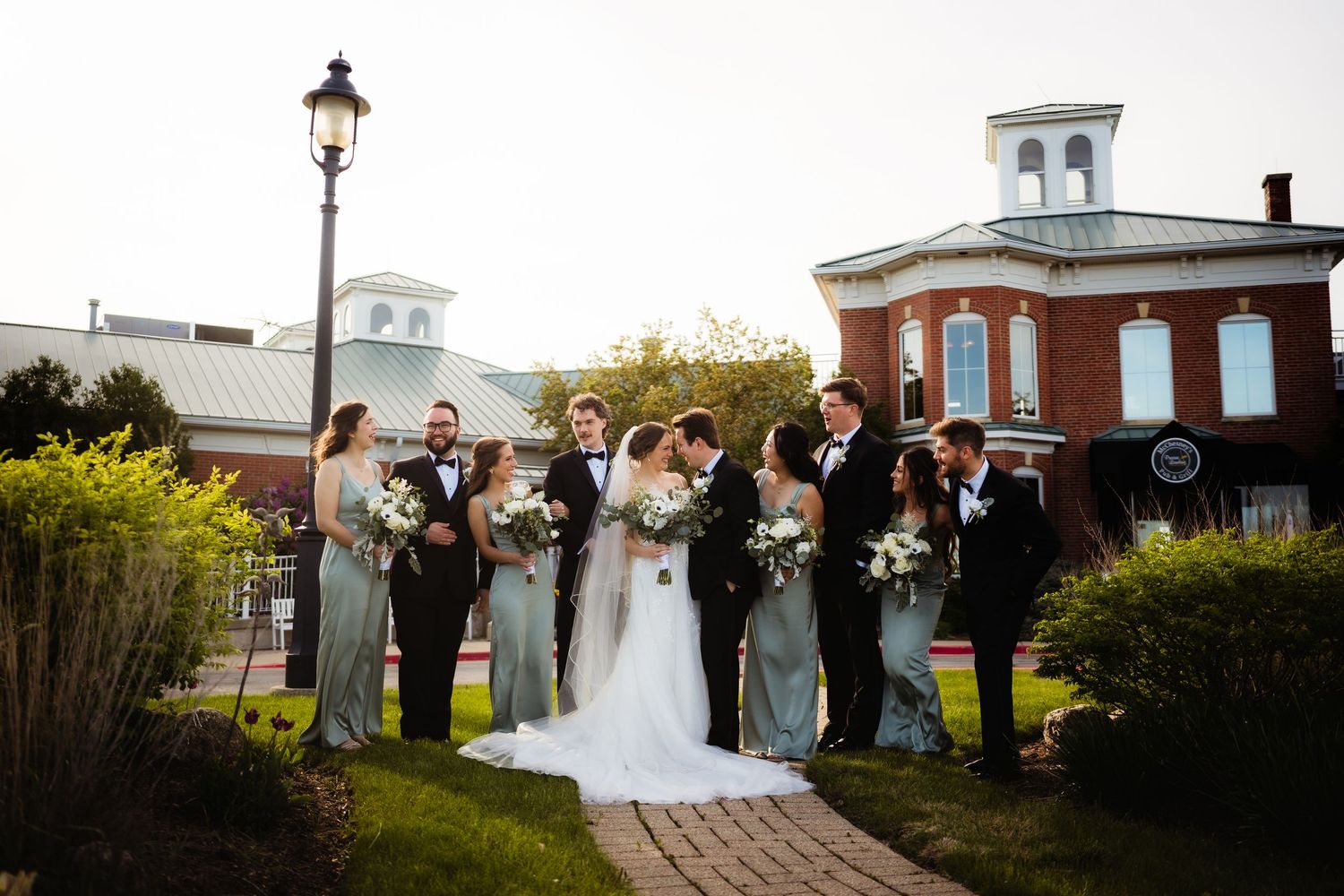 Wedding party in sage green and black tuxedos poses outside a brick venue while bride and groom share a kiss.