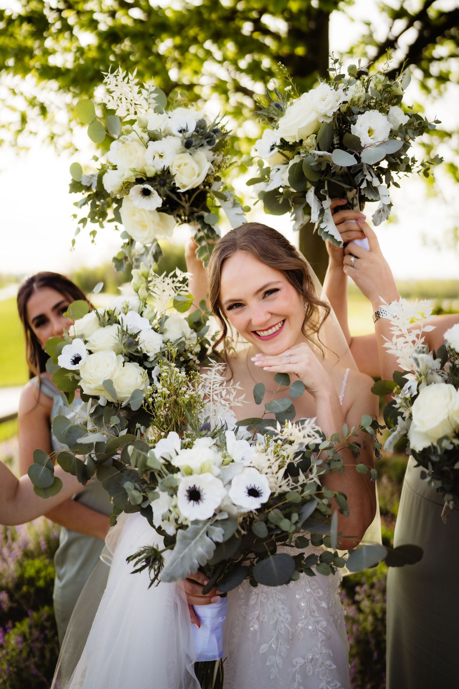 Smiling bride surrounded by bridesmaids holding white floral bouquets with eucalyptus greenery outdoors.