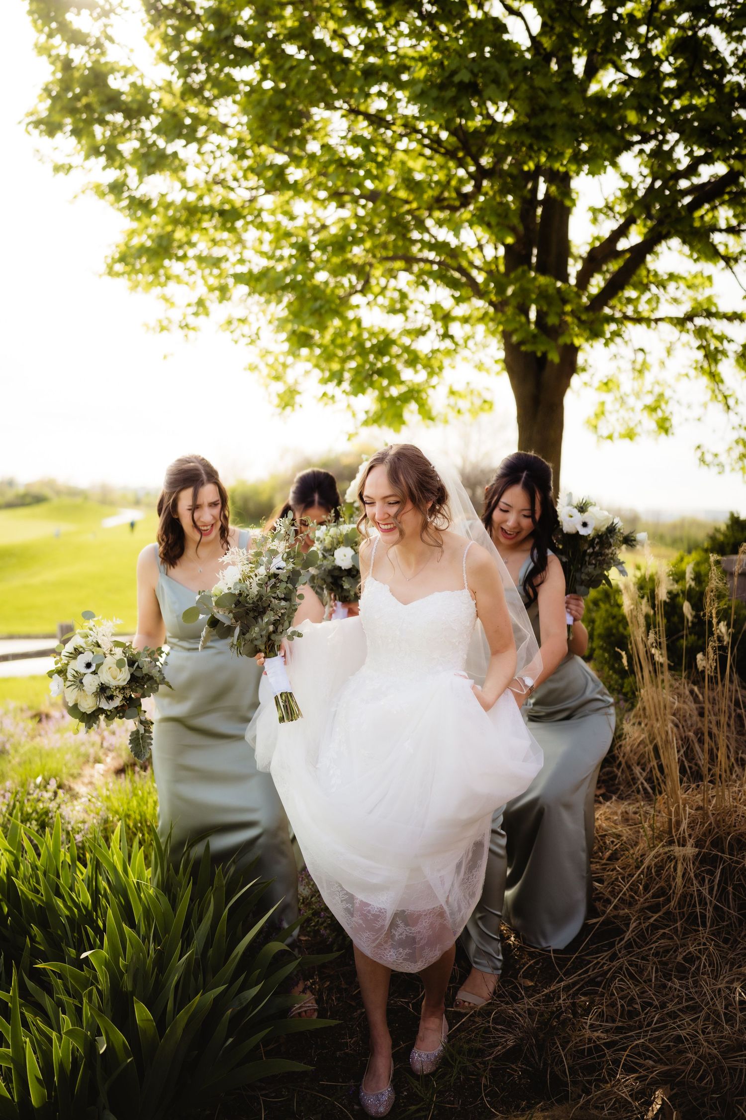 Bride in white gown with bridesmaids in sage green dresses holding bouquets outdoors in golden hour sunlight.