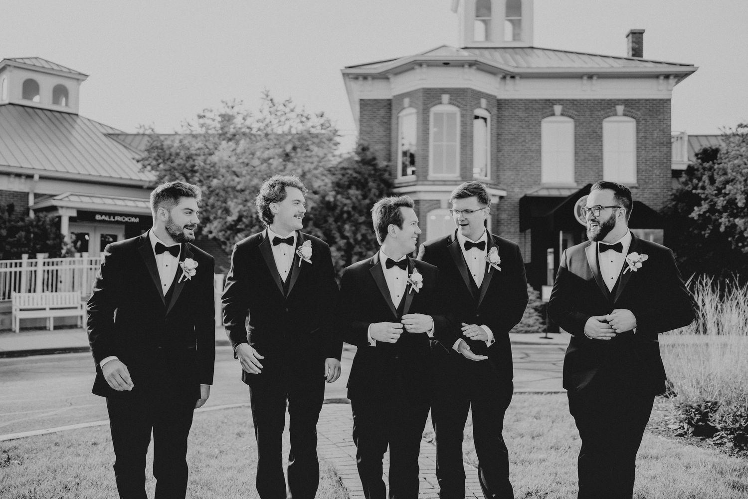 Five groomsmen in black tuxedos and bow ties walk together outdoors near a historic brick building.
