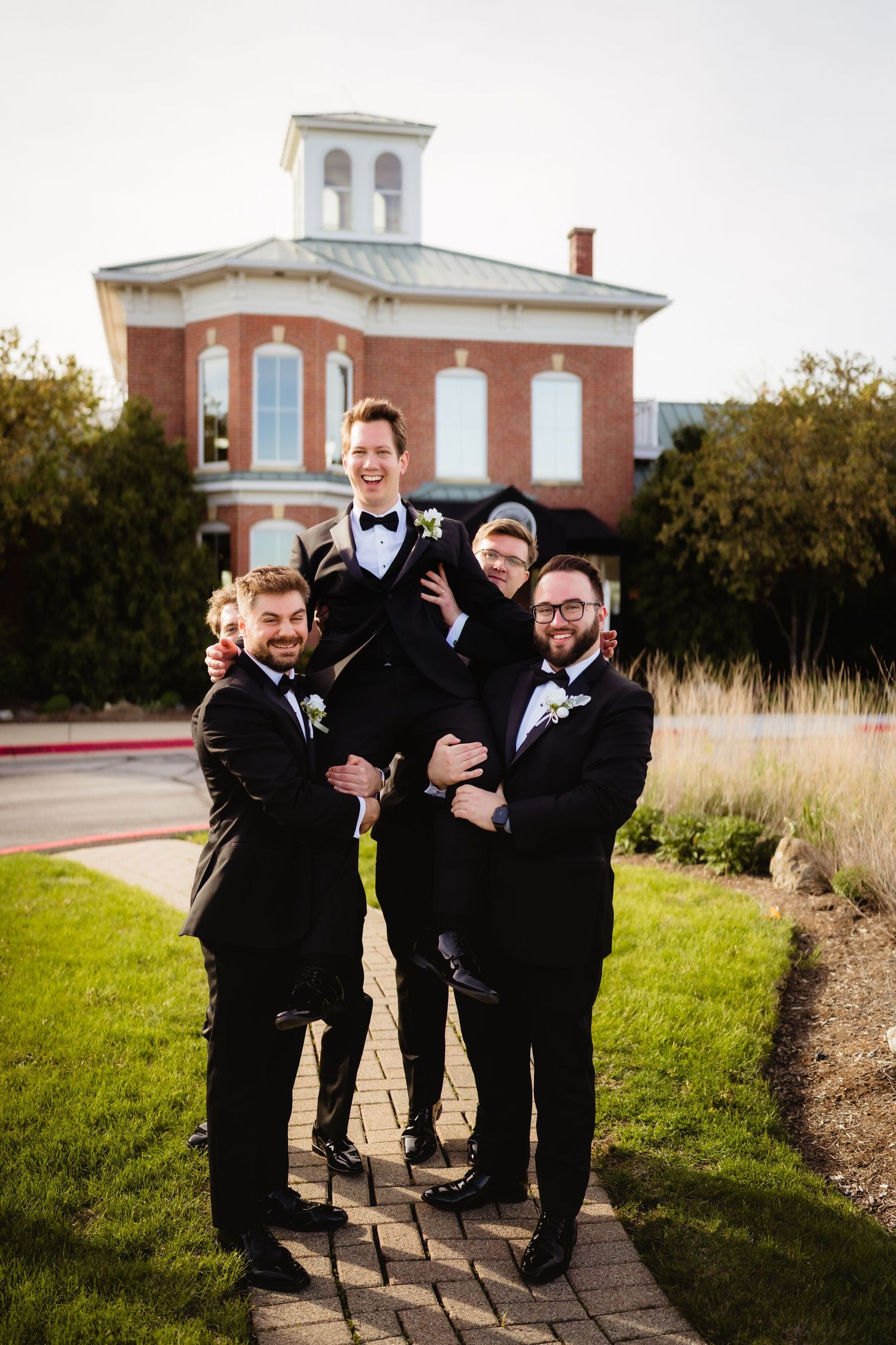 Groomsmen in black tuxedos lifting the groom outside a brick wedding venue on a cloudy day.