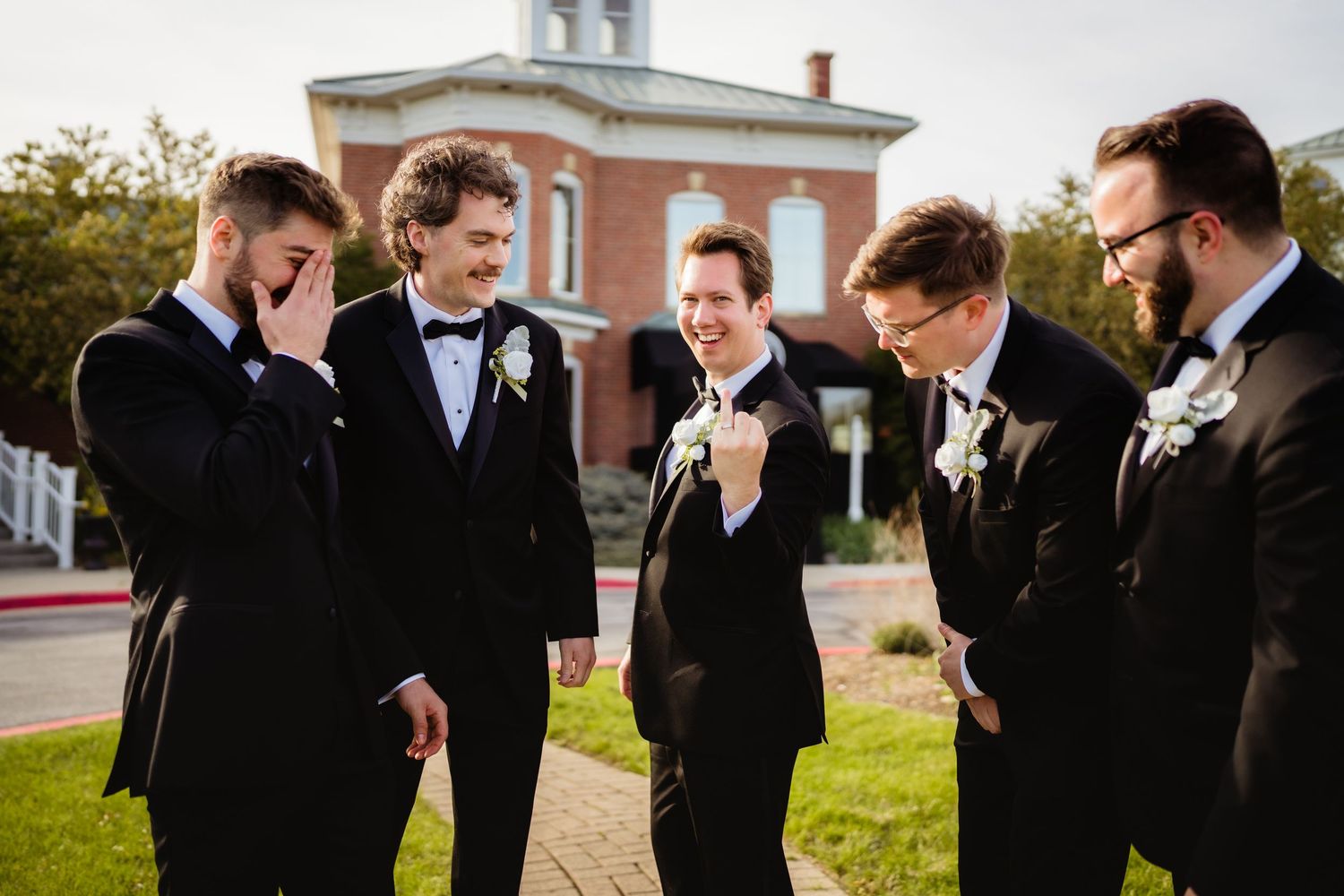 Five groomsmen in black tuxedos laughing together outdoors before a wedding ceremony near a brick building.