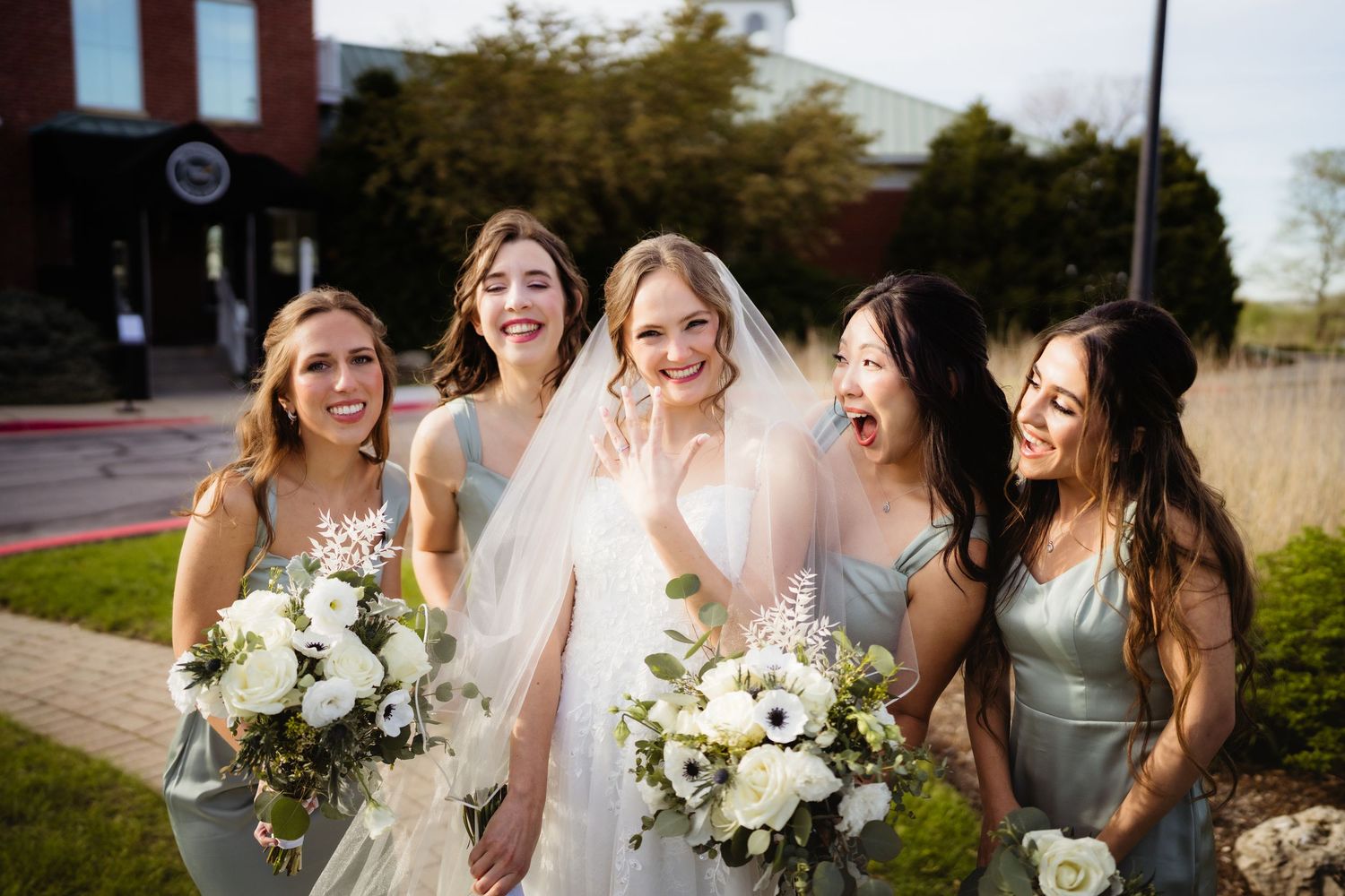 Bride in white gown and veil laughing with four bridesmaids in sage green dresses holding white floral bouquets.