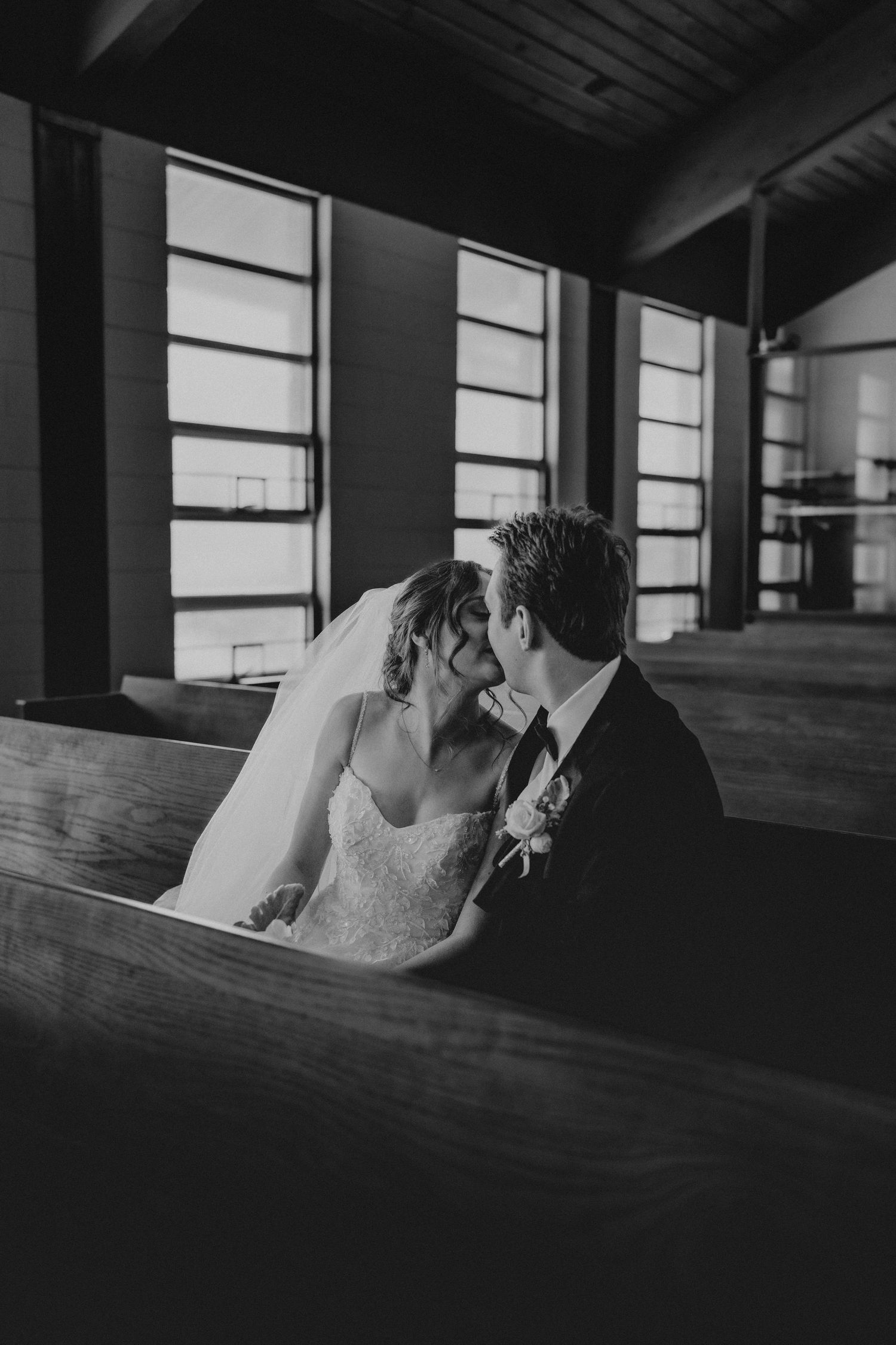 Black and white photo of a bride and groom sharing a kiss on a church pew, with large windows in the background.