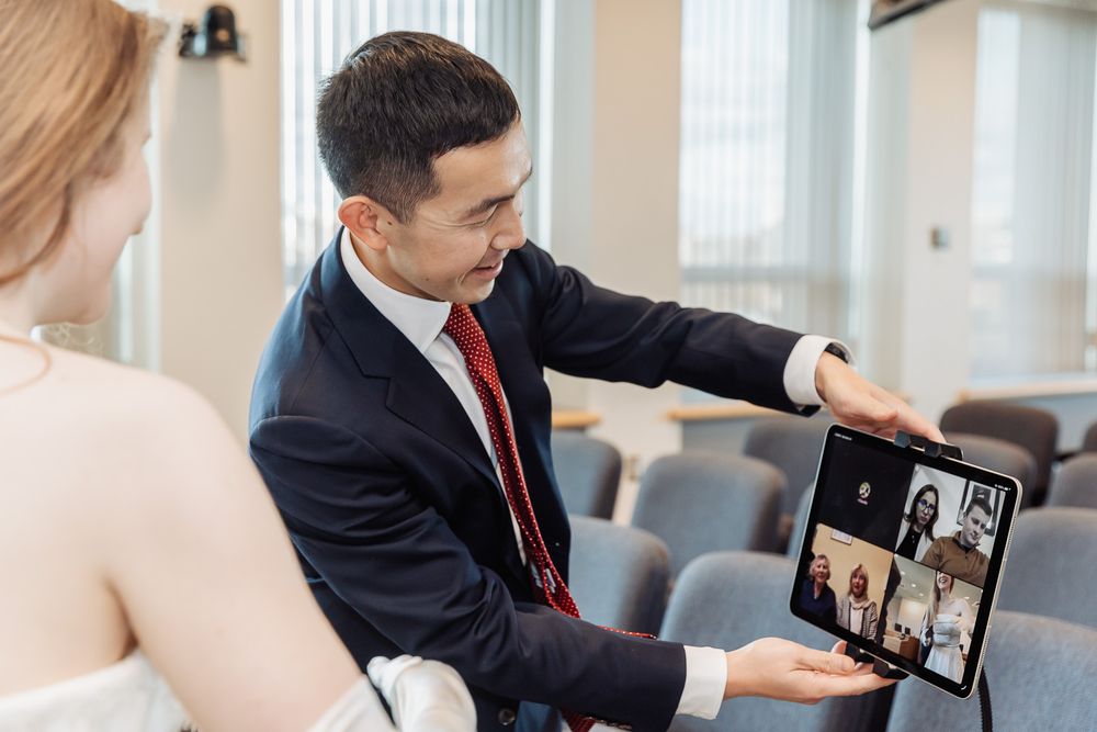 Man in suit shows video call on tablet to woman in white dress during a meeting in a modern conference room.