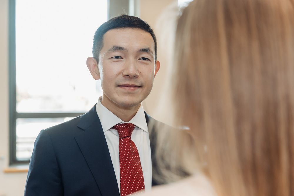 A professional Asian man in a dark suit and red tie smiling during a formal meeting or event.