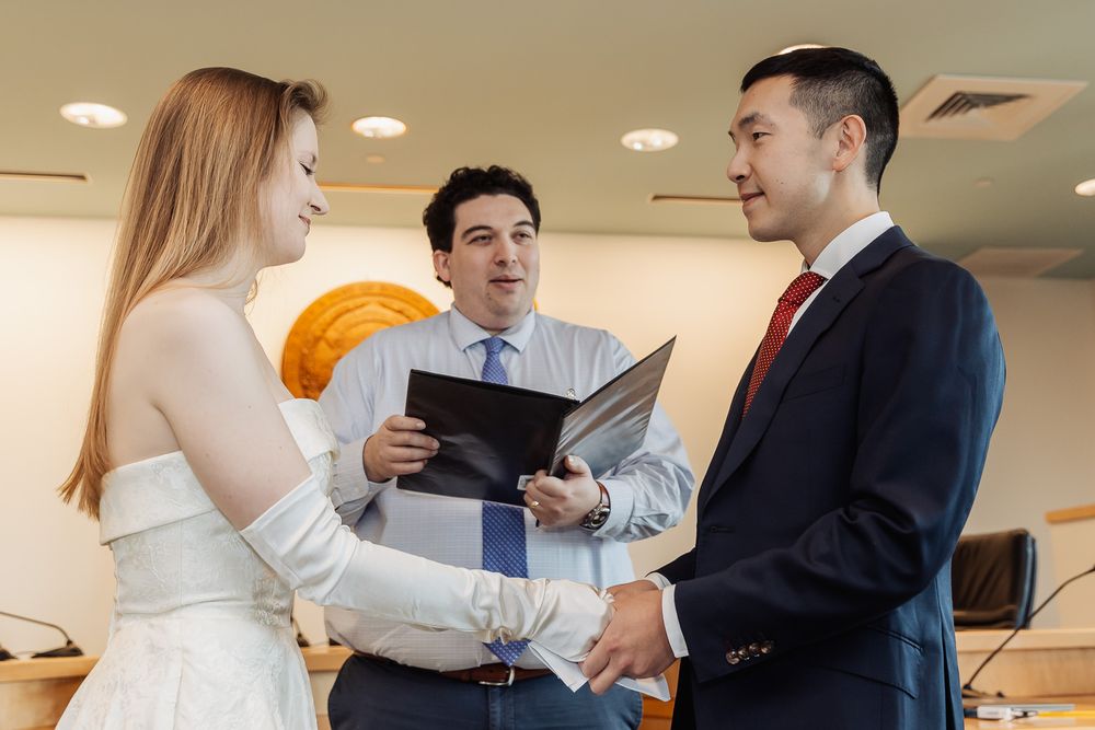 A couple exchanges vows at a civil wedding ceremony while an officiant holds an open binder indoors.