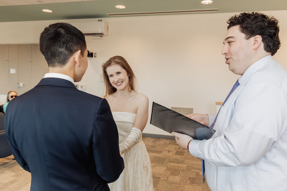 Couple exchanging vows with officiant holding binder during intimate indoor wedding ceremony.