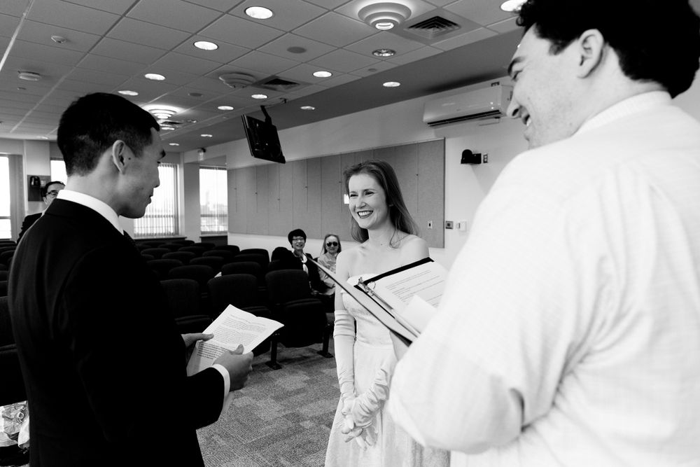 Black and white photo of a couple exchanging vows at a civil wedding ceremony indoors with an officiant.