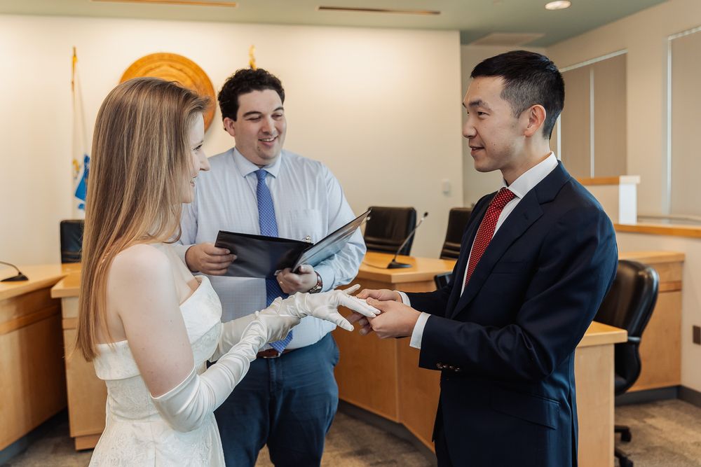 Couple exchanging rings during a civil courthouse wedding ceremony with an officiant holding a folder.