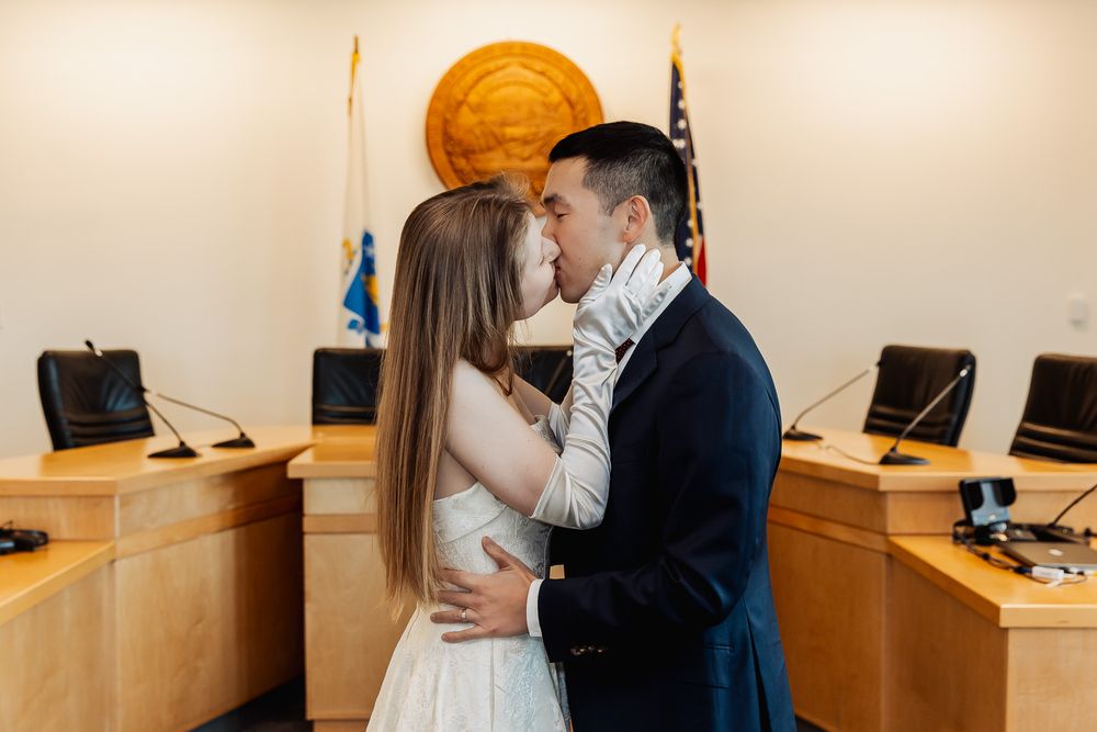 Couple sharing a first kiss as newlyweds in a courthouse ceremony room with American flag backdrop.