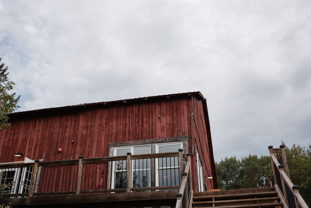Red barn with wooden deck and staircase against cloudy sky, surrounded by trees.