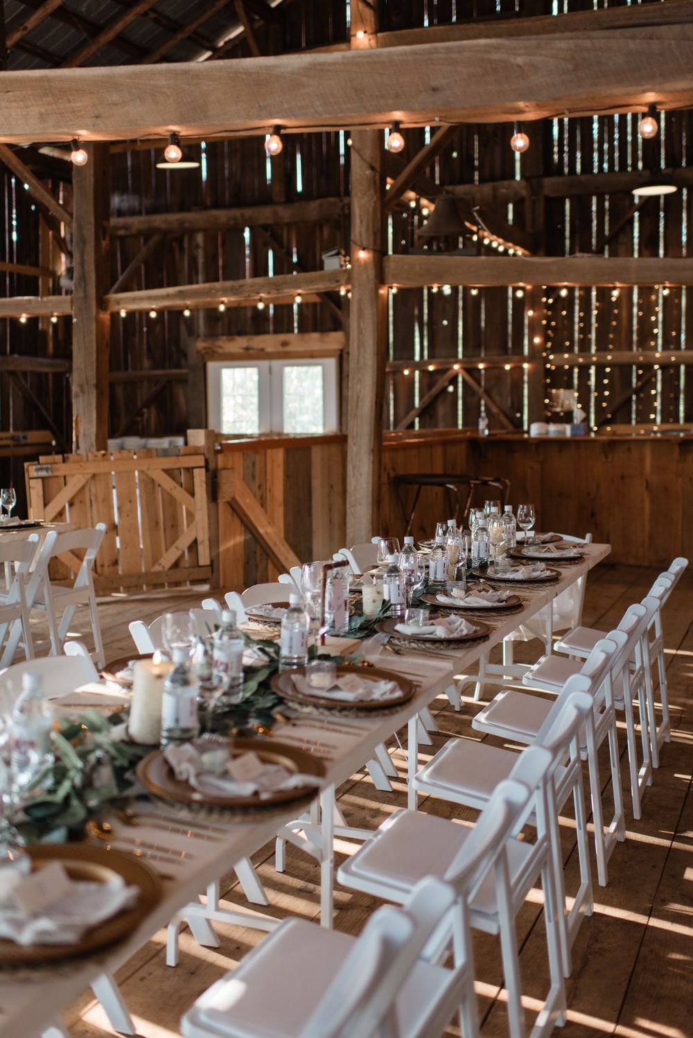 Rustic barn wedding reception with long wooden tables, white chairs, string lights, and elegant greenery centerpieces.
