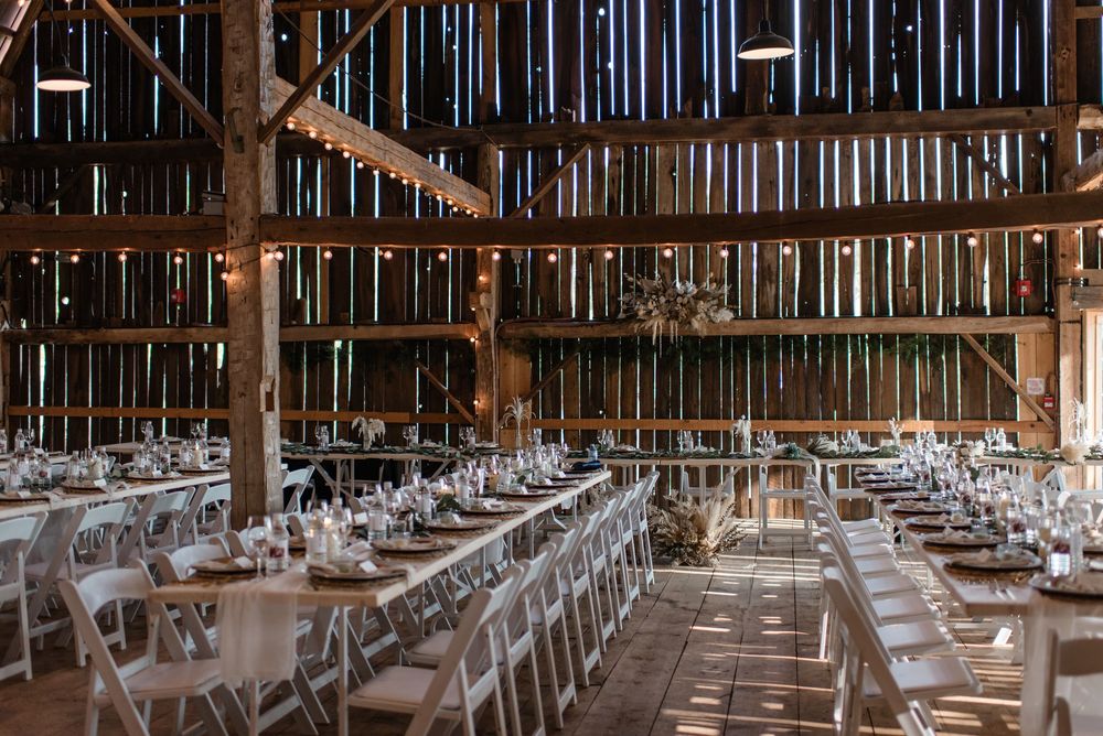 Rustic barn wedding reception with long tables, white chairs, string lights, and wooden beamed ceilings.