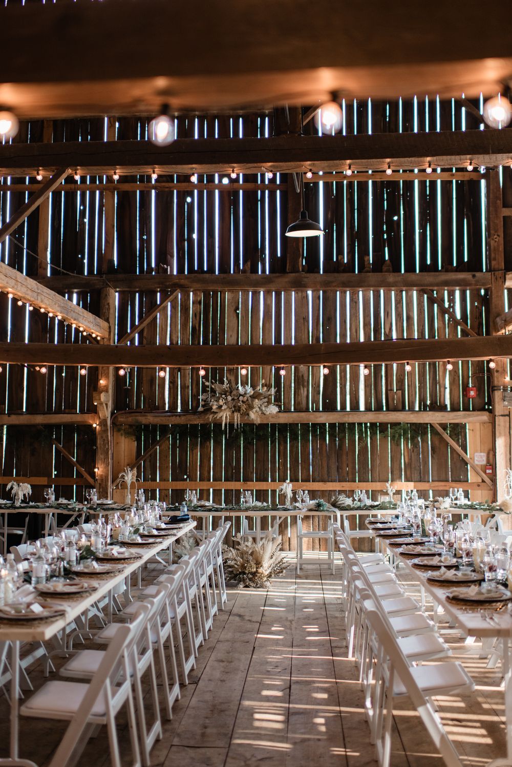 Rustic barn wedding reception with long white tables, string lights, and wooden beamed ceiling.