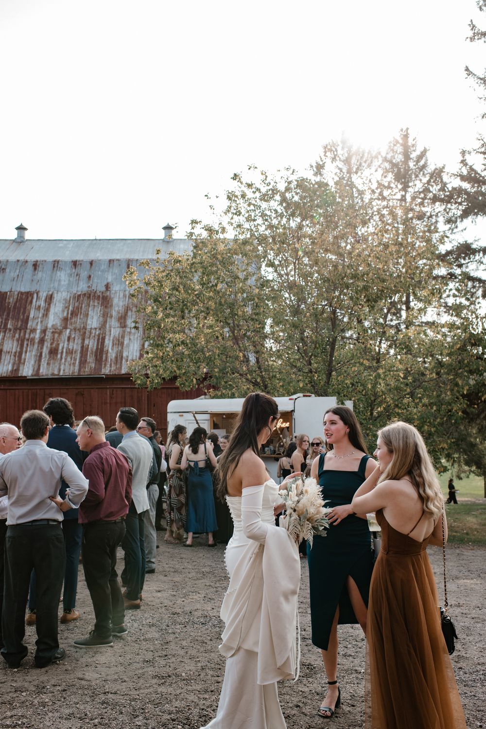 Bride in white off-shoulder gown holds bouquet chatting with guests at rustic outdoor wedding reception near barn.