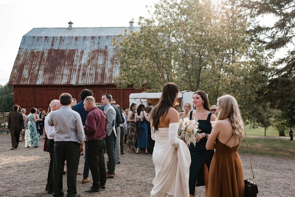 Guests mingle outdoors at a rustic barn wedding venue, with bride in white gown holding bouquet.